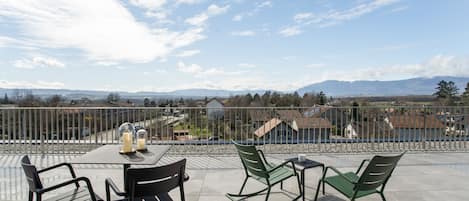 A terrace with a table and chairs, a view of houses and mountains in the distance, and a clear sky.