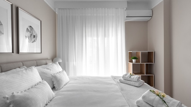 Ein modernes Hotelzimmer mit einem großen Bett, einem Nachttisch, einer kleinen Holzwandregal mit einer Pflanze und einem großen Fenster mit durchsichtigen Vorhängen.