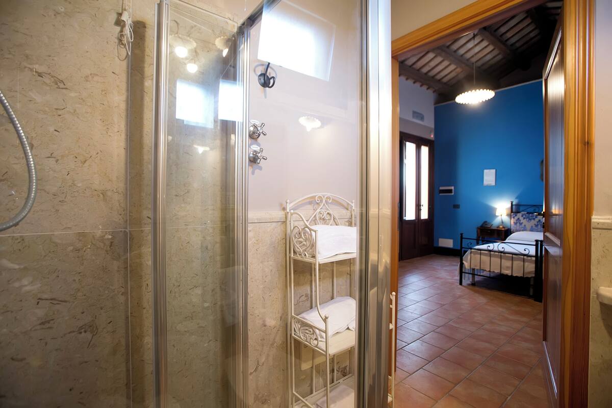 Een badkamer met een douchecabine van glas, een witte metalen plank en een bed in de aangrenzende kamer.