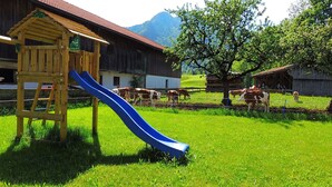 Un terrain de jeux avec un toboggan et une maisonnette en bois, le tout situé devant une grange et un champ où paissent des vaches.