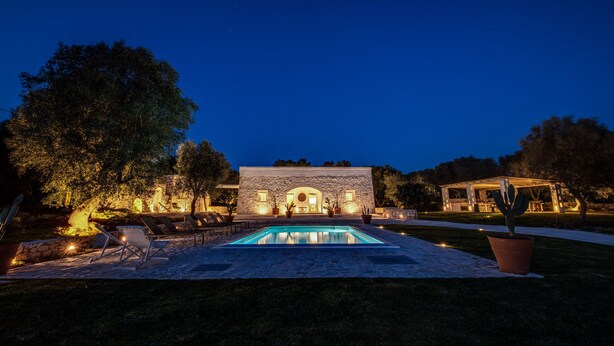 A pool with lounge chairs, a stone building with a circular window, and a pergola with lights.