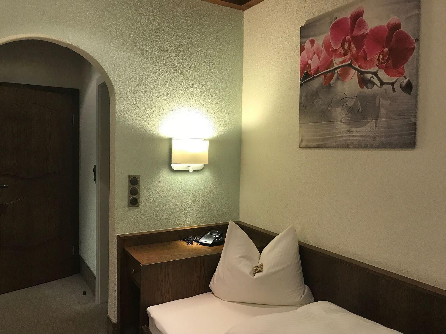 Habitación de hotel con cama, mesita de noche, lámpara, un cuadro de flores y una puerta.