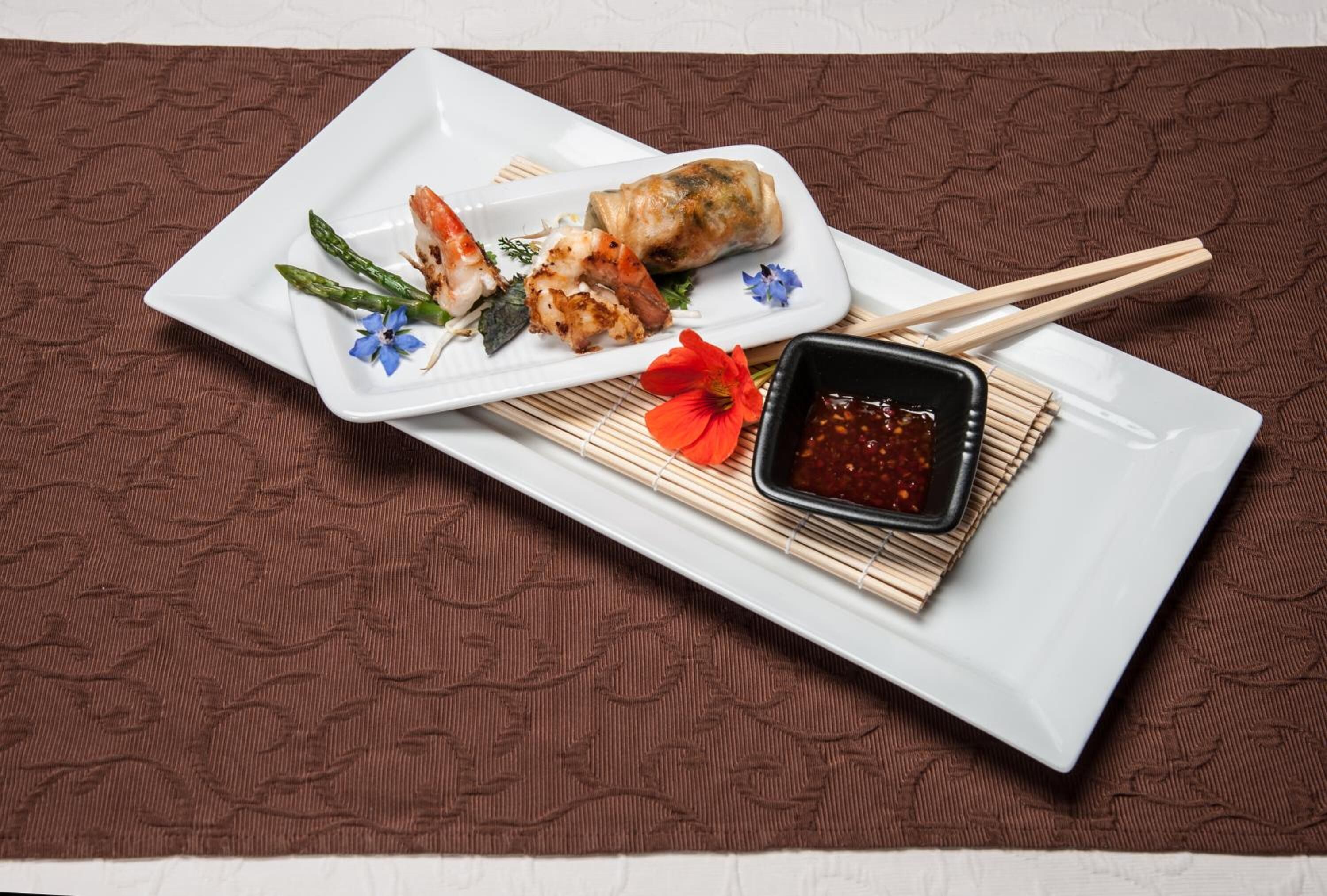 A white rectangular plate with a spring roll, asparagus, and a flower, accompanied by a small bowl of sauce and chopsticks on a brown textured mat.
