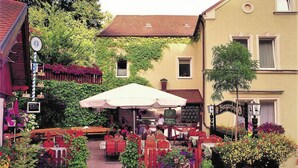 Un café en plein air avec des tables et des chaises, entouré de verdure et bordé par un bâtiment sur lequel on peut voir une enseigne.