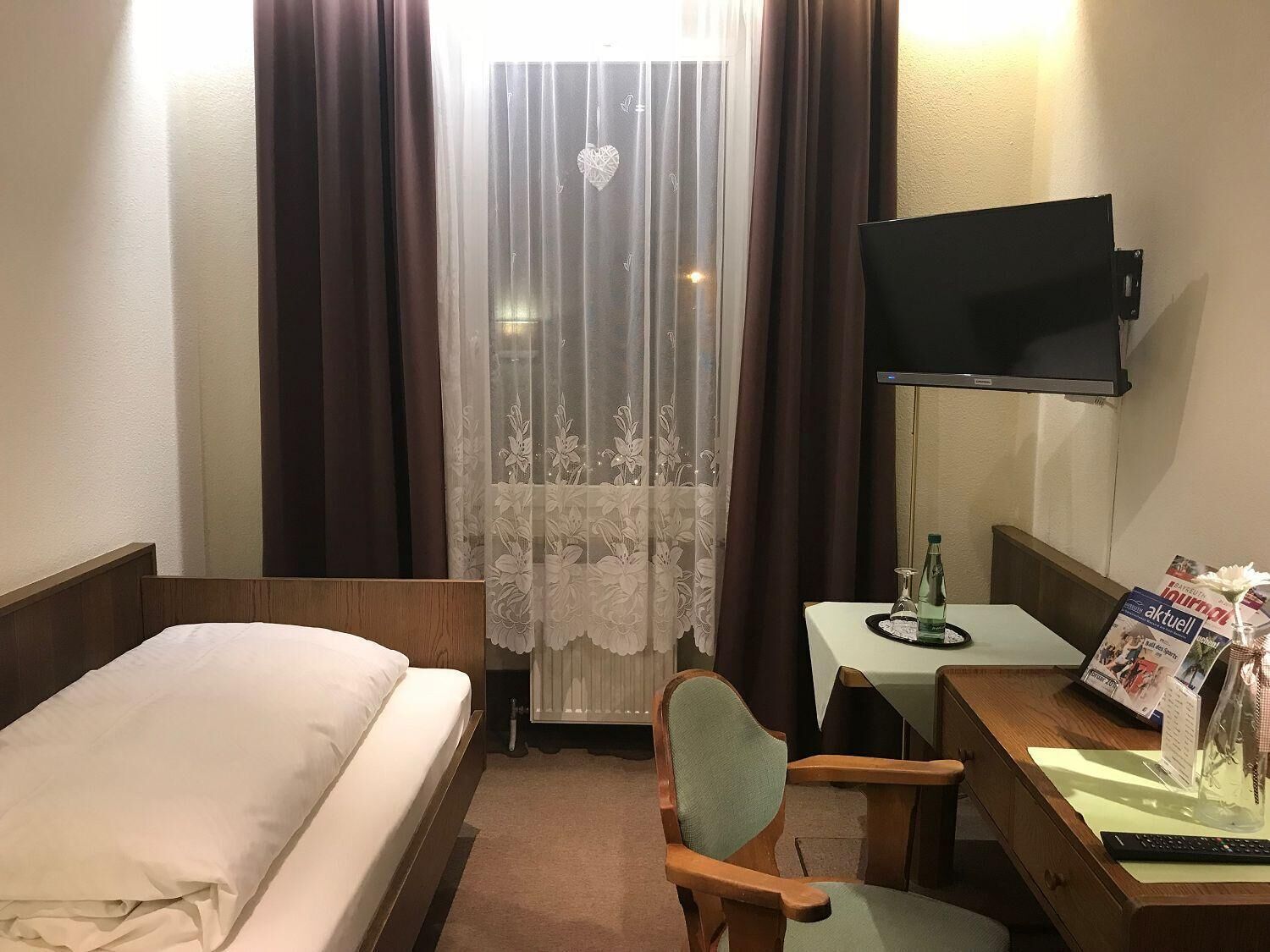 Ein Hotelzimmer mit einem Bett, einem Schreibtisch mit Stuhl, einem Fernseher und einem Fenster mit Vorhängen.