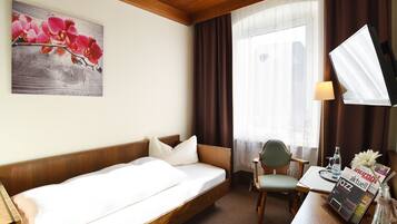 A hotel room with a bed, a desk with a chair, a lamp, a TV, and a window with curtains.