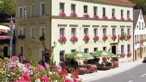 Un bâtiment à deux étages, doté d’un toit plat et de nombreuses fenêtres, dont certaines sont ornées de jardinières. On y trouve des tables à l’extérieur, protégées par des parasols.