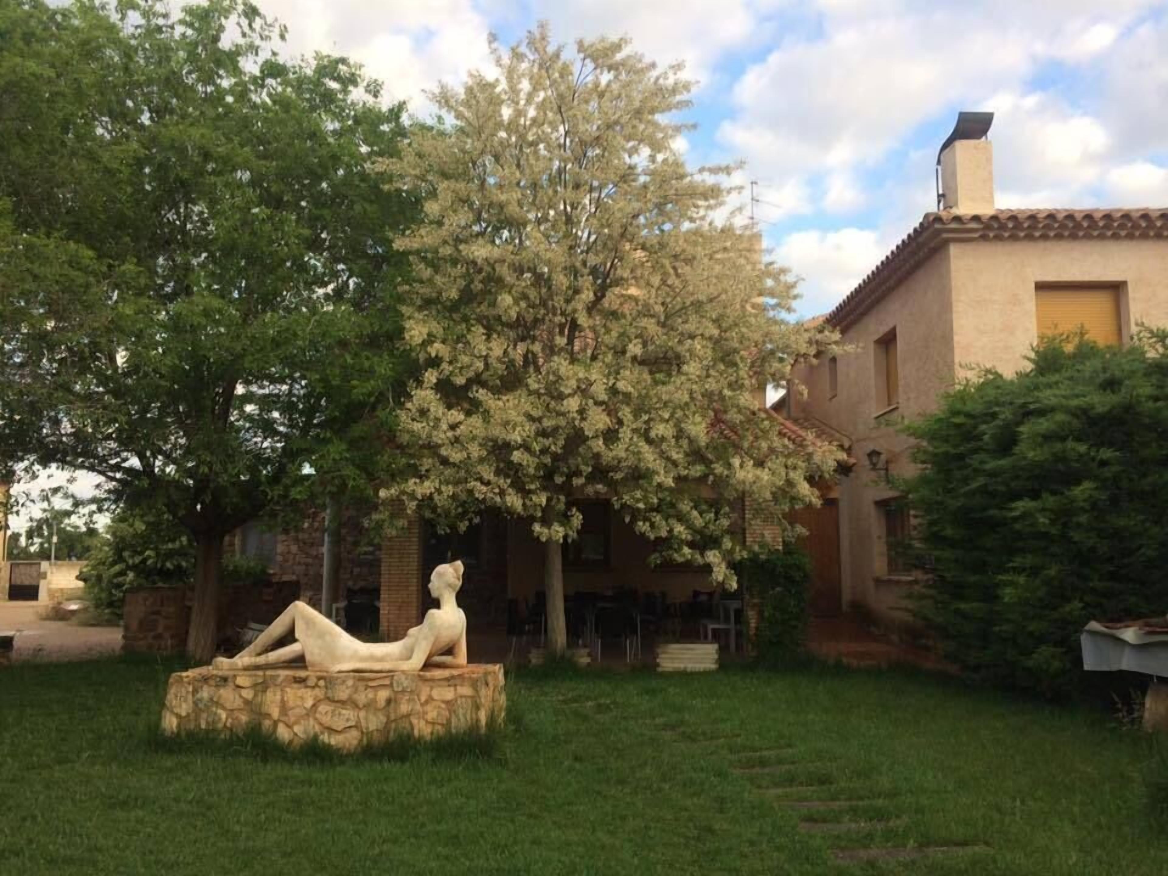Una escultura de una figura reclinada en una cuenca de piedra, rodeada de exuberante follaje y un edificio con tejado de azulejos.