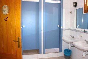 A bathroom with a wooden door, a blue shower enclosure, two sinks, and a trash bin.