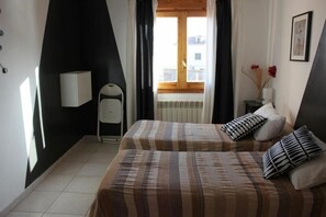A bedroom with two beds, a window with curtains, a bedside table with a vase of flowers, and a wall-mounted lamp.