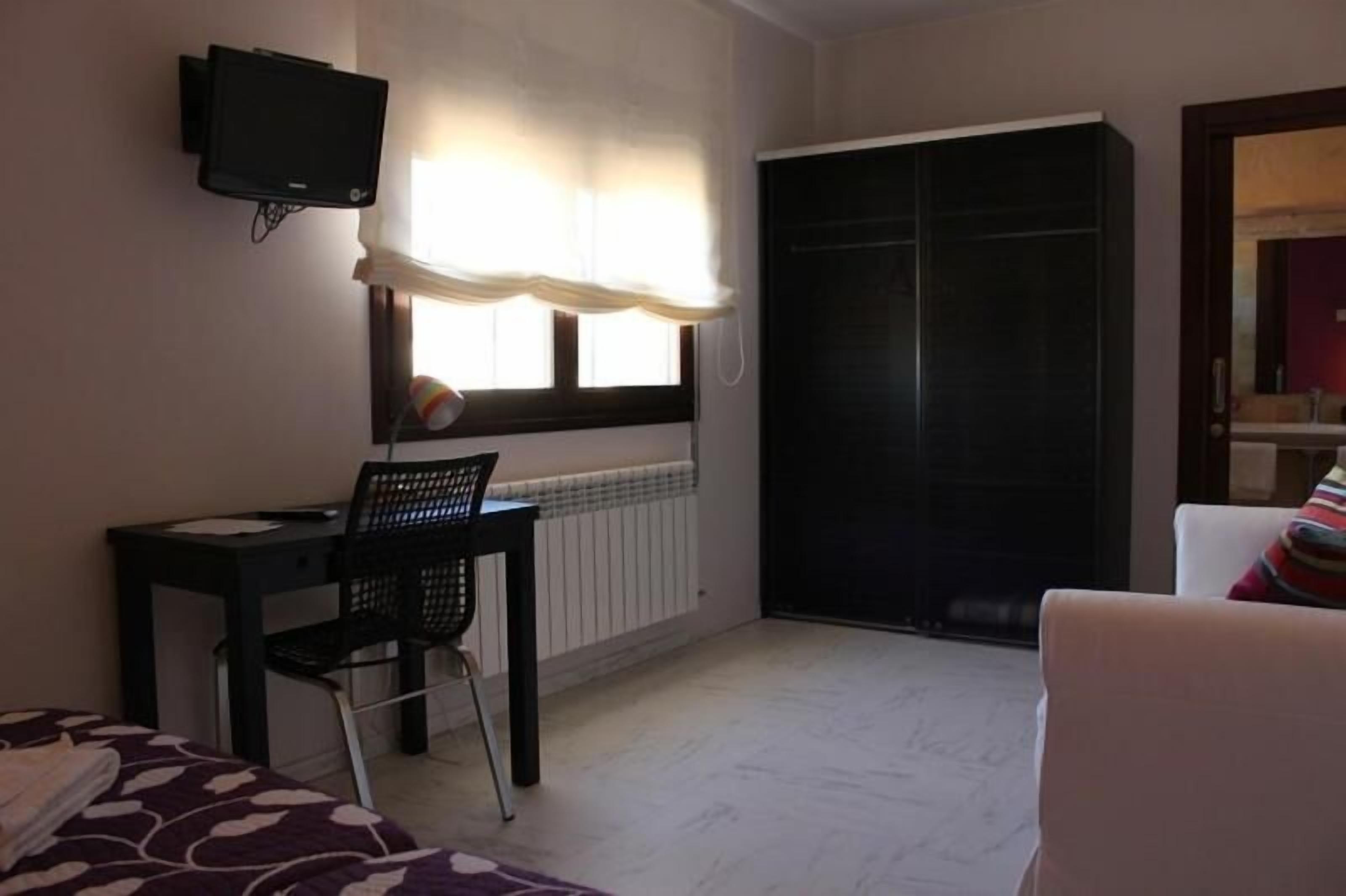 Habitación de hotel con cama, escritorio y silla, televisor montado en la pared, ventana con cortinas, armario y puerta.