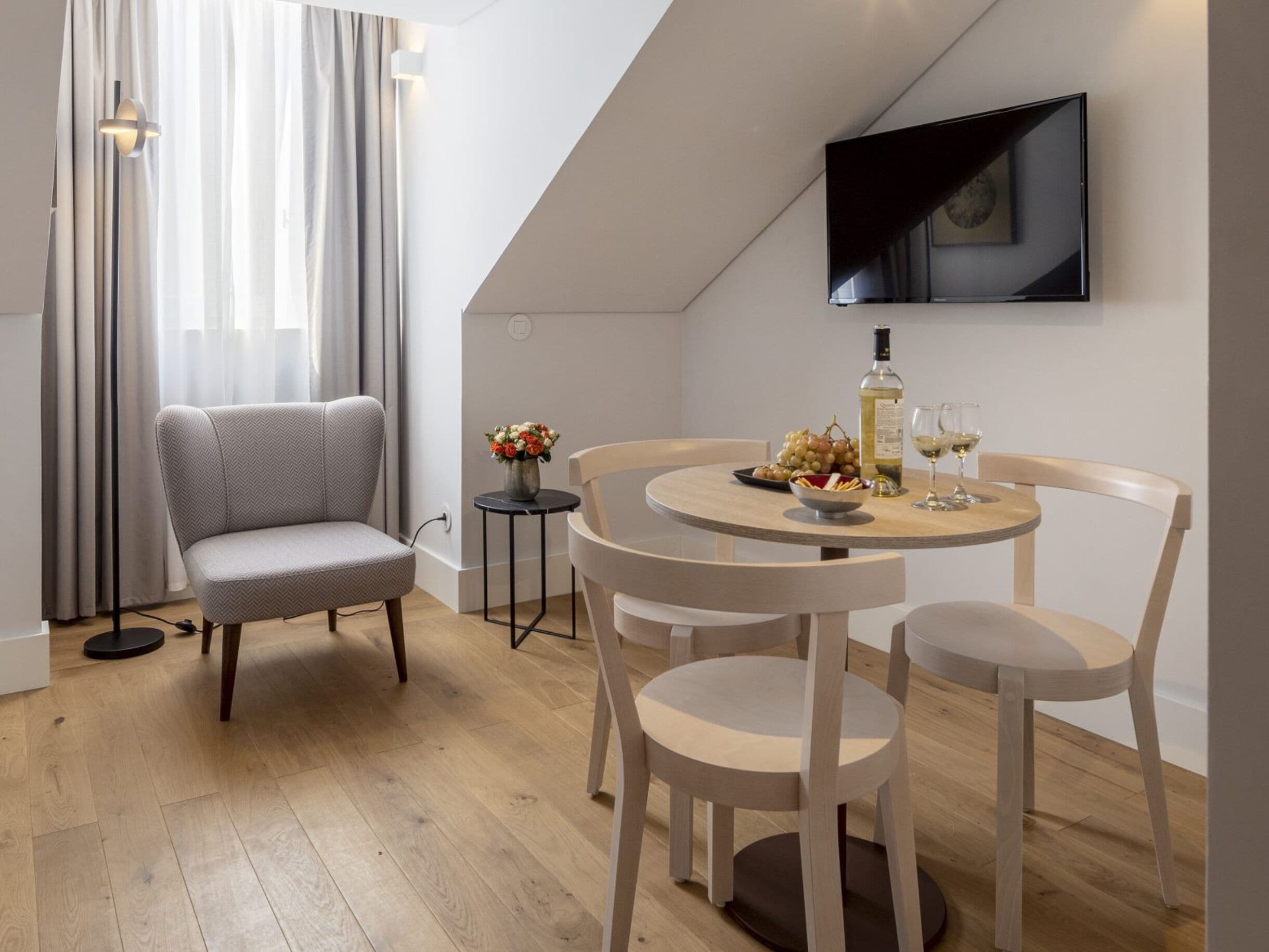 A modern living room with a grey sofa, a round wooden dining table, and a flat-screen TV mounted on the wall.