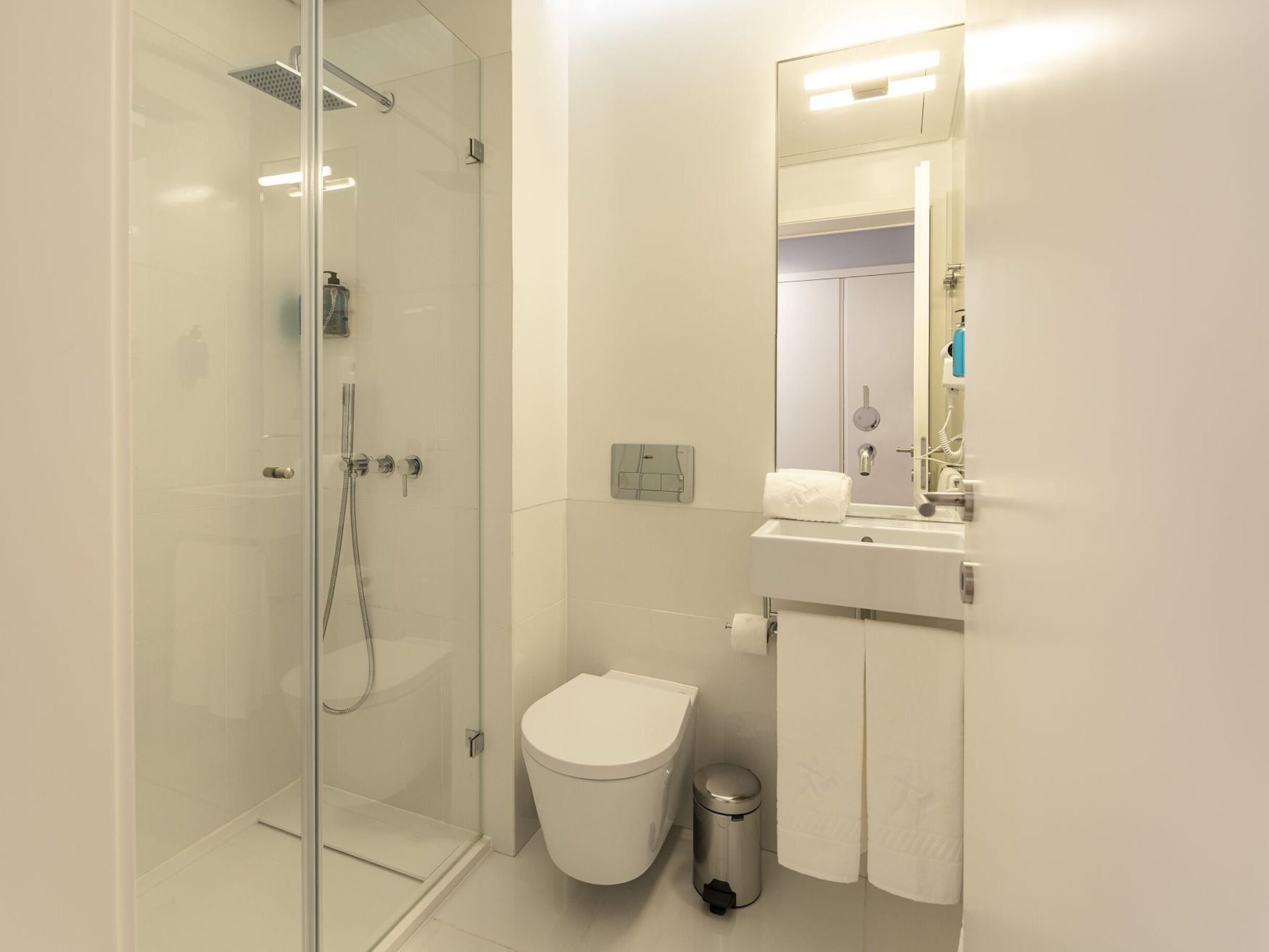 A modern bathroom with a glass-enclosed shower, a white toilet, a sink with a mirror, and a trash bin.