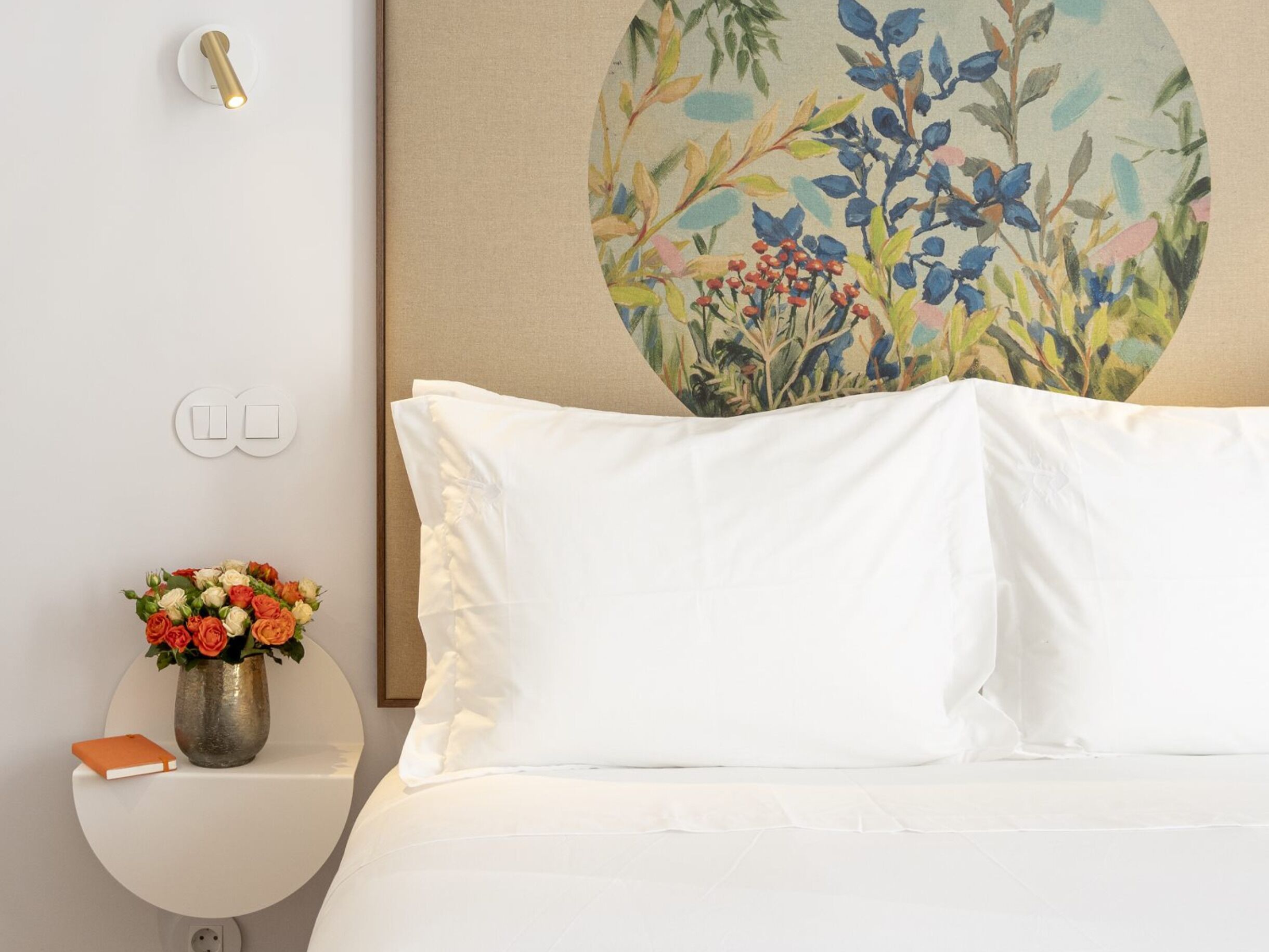 A neatly made bed with white bedding, a wall-mounted light fixture, a round mirror, and a floral painting.