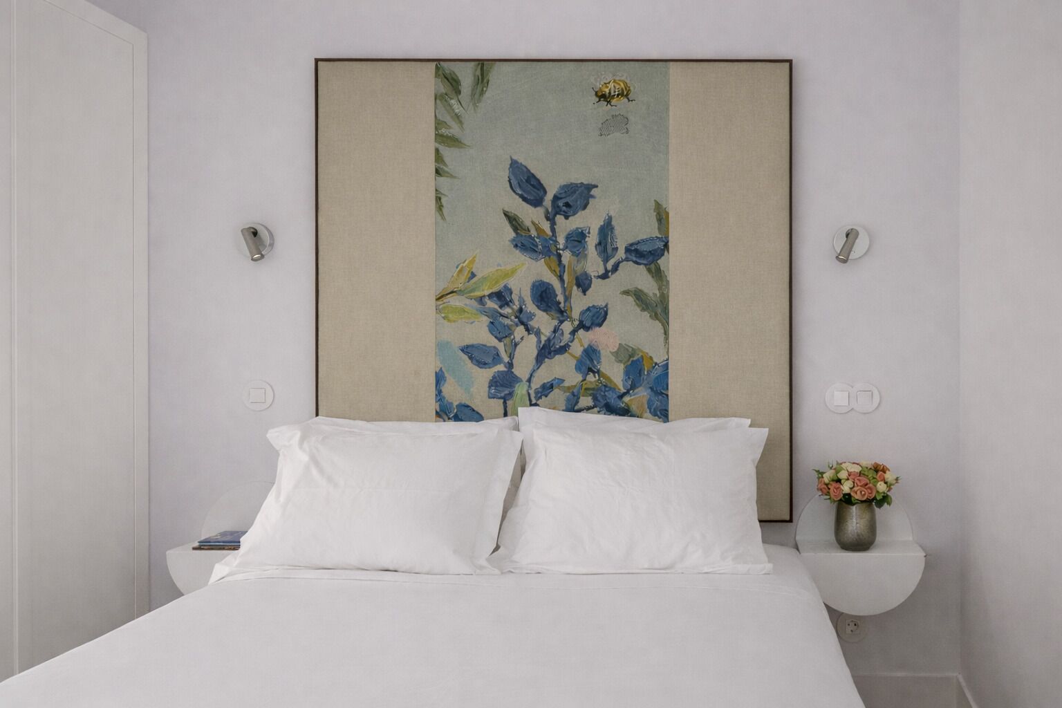 A bed with white linens, a nightstand with a book and a vase of flowers, and a wall-mounted painting with blue and green foliage.