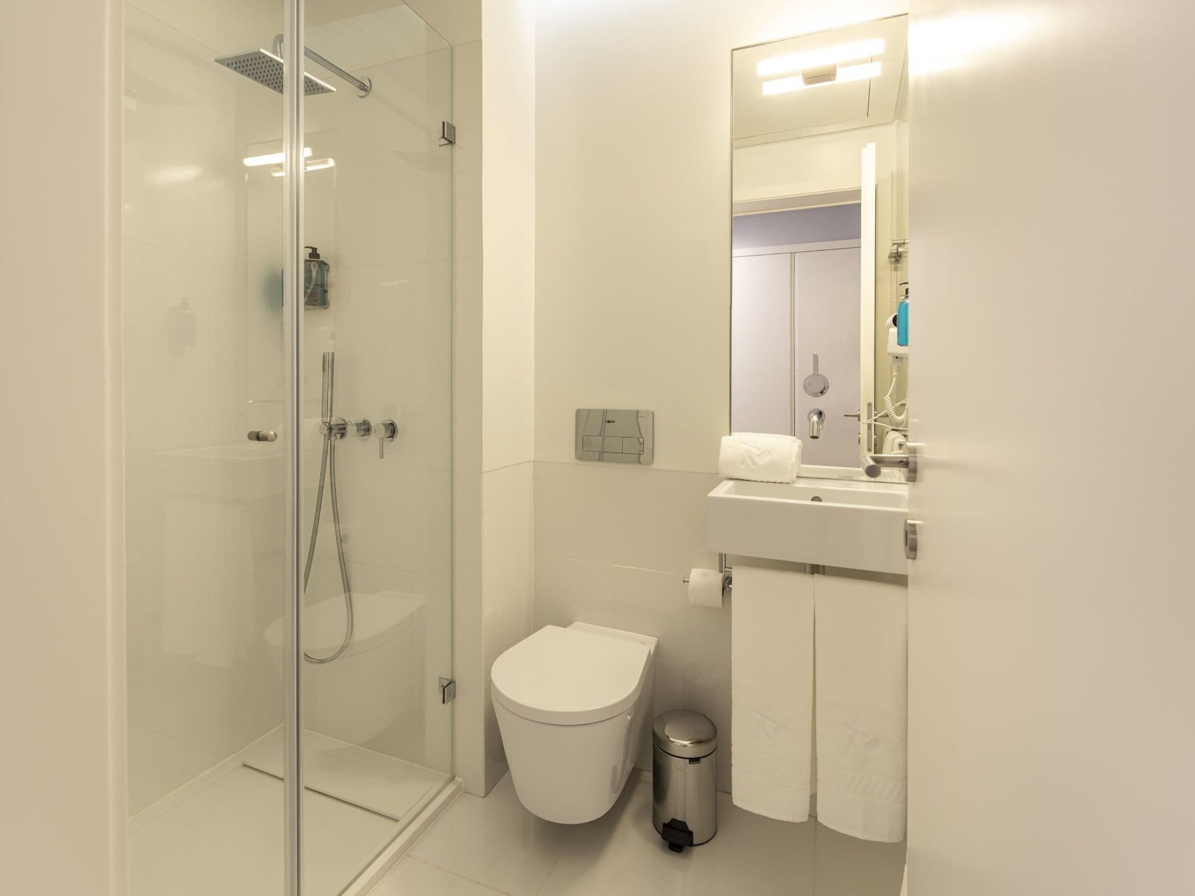 Un bany modern amb plat de dutxa de vidre, vàter blanc, paperera i un lavabo amb mirall.