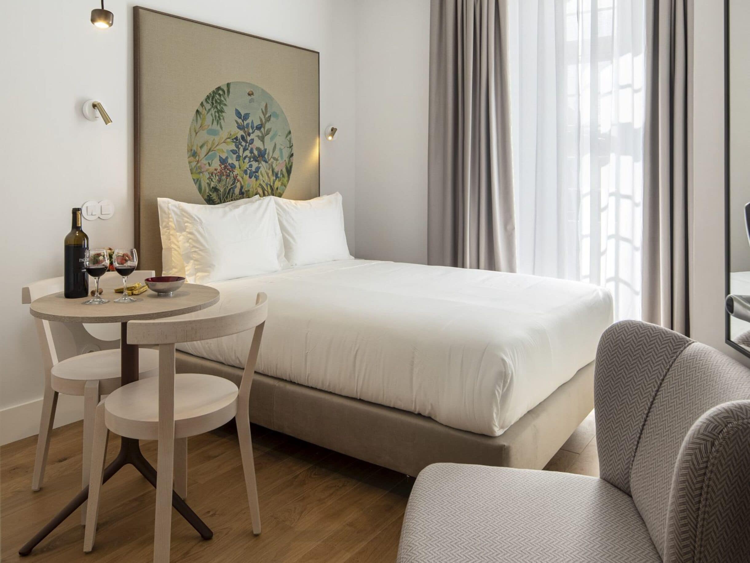 Une chambre d’hôtel avec un lit, une table ronde entourée de chaises, une bouteille de vin et un tableau accroché au mur.