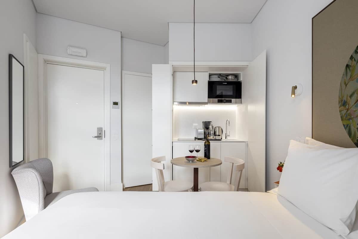 A hotel room with a bed, a small dining table with chairs, a kitchenette, and a single hanging light fixture.