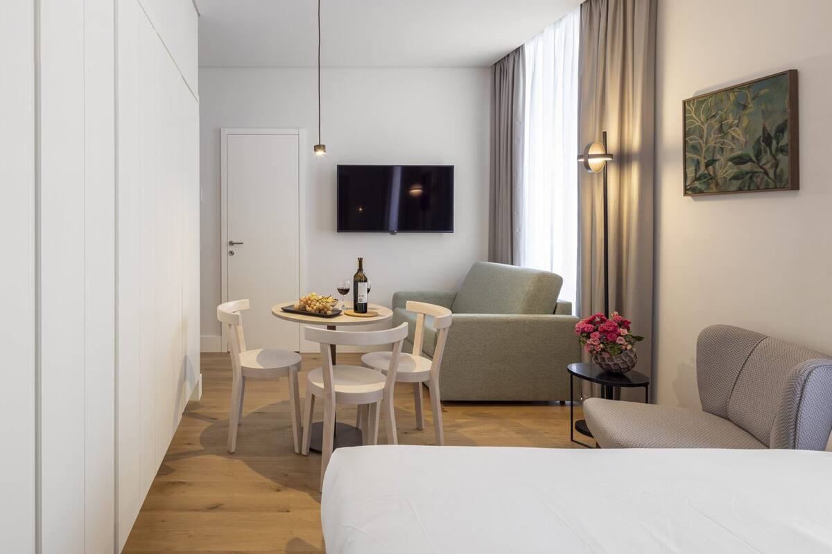 A modern hotel room with a bed, a dining area with chairs and a table, a sofa, and a TV mounted on the wall.