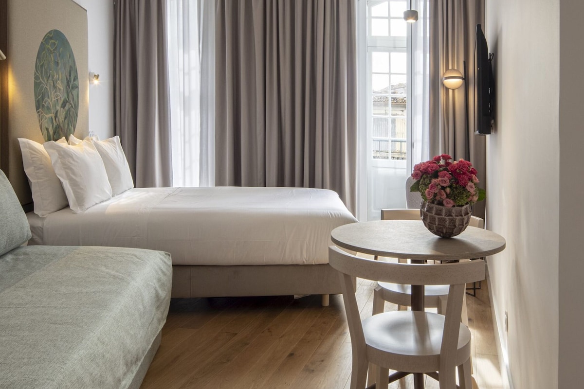 A hotel room with a bed, a seating area with a table, and a large window with curtains.