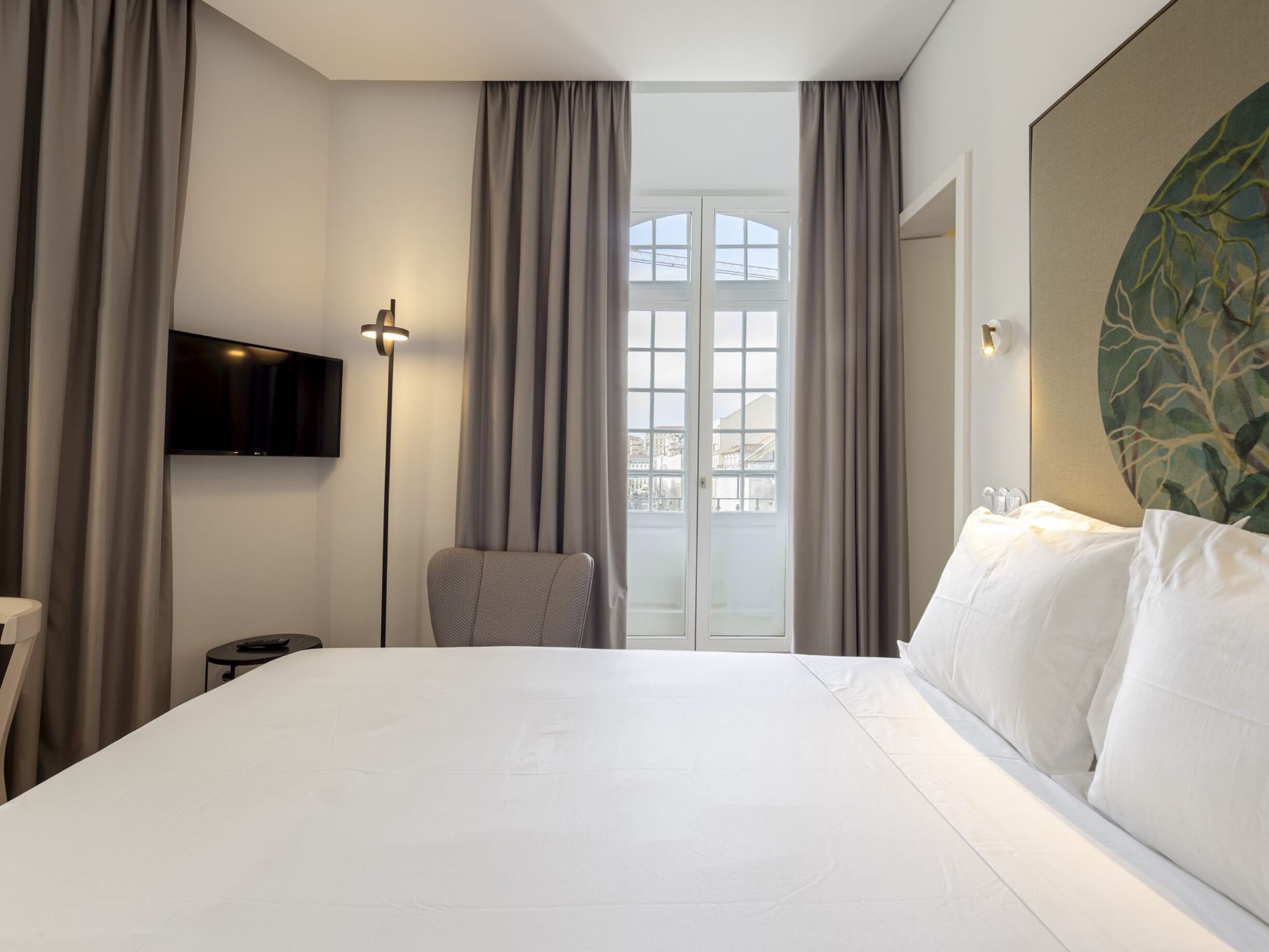A hotel room with a large bed, a chair, a TV, and a view of the city through a window.