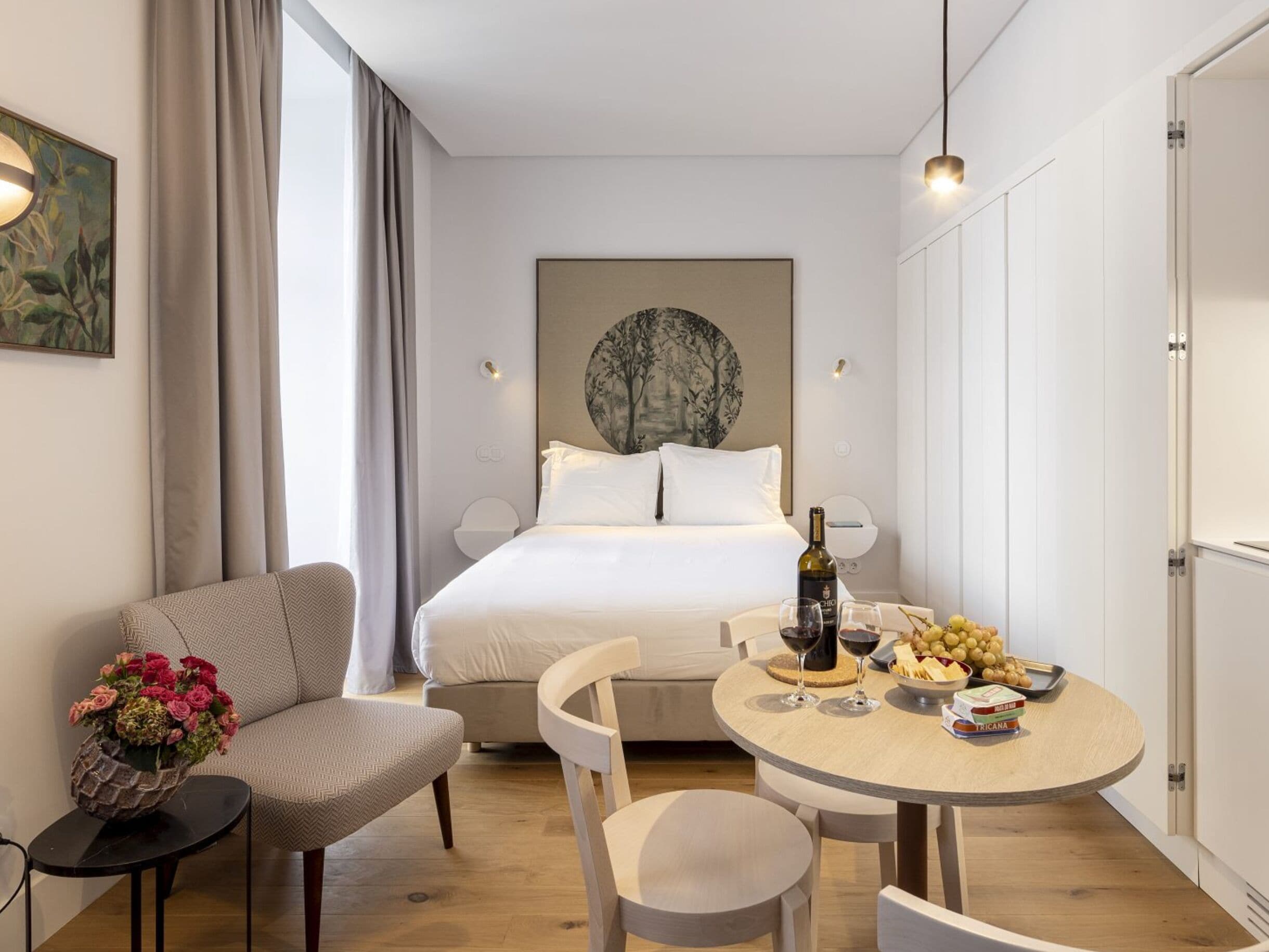 Una habitación de hotel moderna con una cama, una mesa de comedor con sillas, una mesita auxiliar con un jarrón de flores y una obra de arte colgada en la pared.