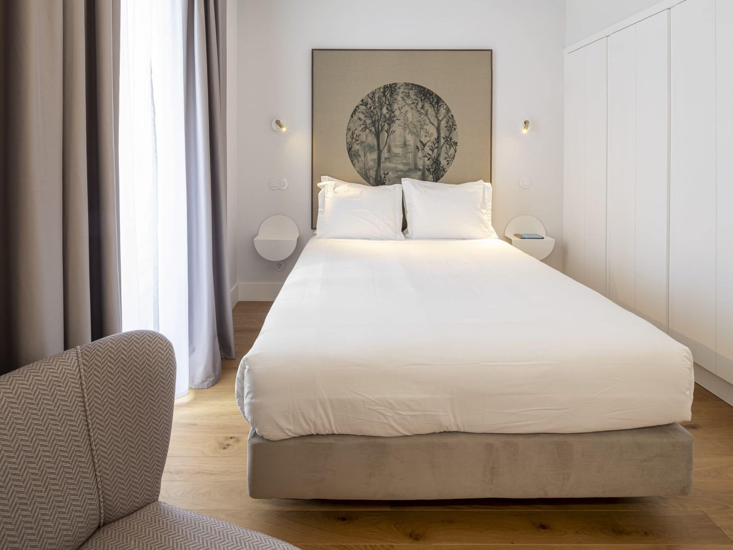 A modern bedroom with a large bed, a side table, a wall-mounted lamp, and a painting above the bed.