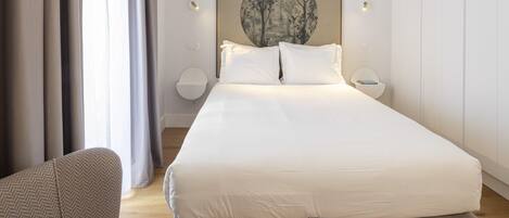 A modern bedroom with a large bed, a side table, a wall-mounted lamp, and a painting above the bed.