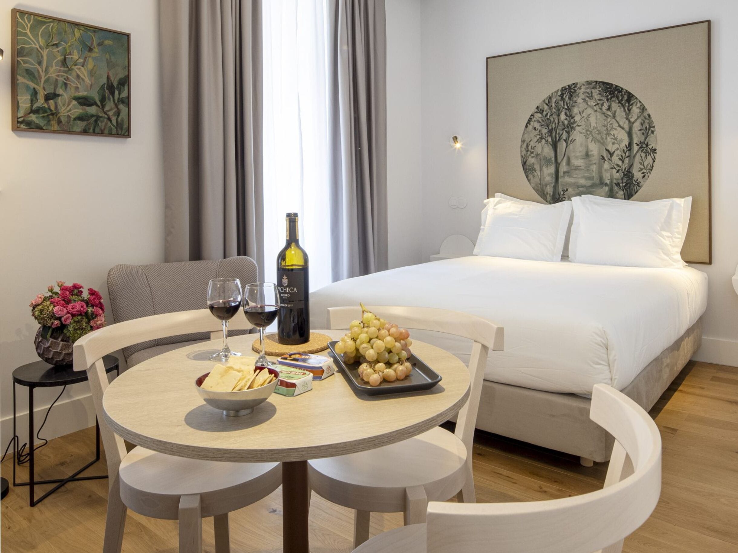 Quarto de hotel com uma cama, uma mesa de jantar redonda com cadeiras, uma garrafa de vinho e uma tigela de uvas.