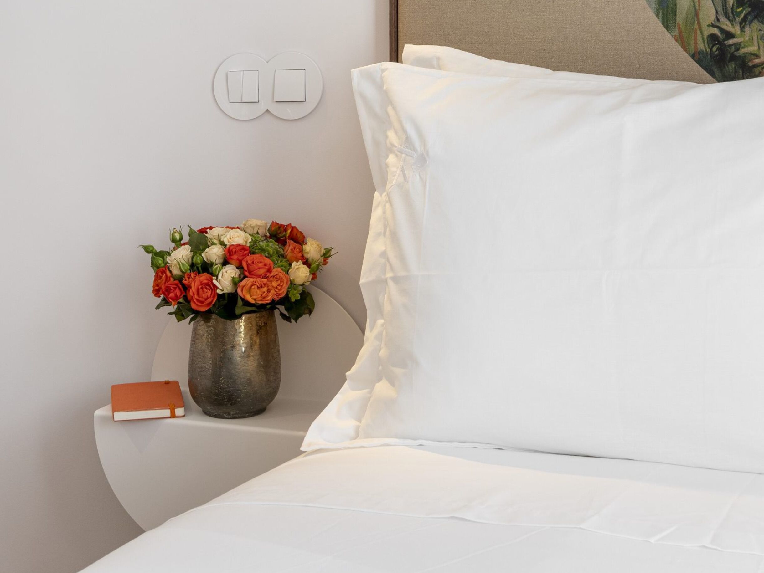 Una cama bien hecha con sábanas blancas, un almohadón blanco y una mesita de noche con un florero de metal y flores.