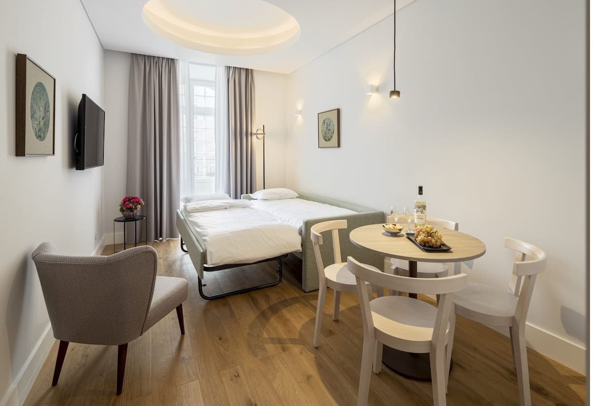 A modern hotel room with a bed, a dining table with chairs, a TV, and artwork on the walls.