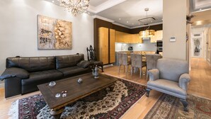 A living room with a dark leather sofa, a wooden coffee table, and a patterned rug. There is a gray armchair and a chandelier. A kitchen is visible in the background. - Aristotelous Grand View (Thessaloniki)