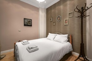 Una cama bien hecha con sábanas blancas y toallas dobladas, piso de madera y una pared con cuadros enmarcados y un perchero.