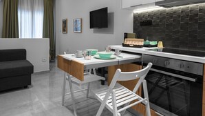 Eine moderne Küche mit Einbauofen, ein Essbereich mit weißem Tisch und Stühlen sowie ein graues Sofa in einem Wohnbereich mit Fernseher an der Wand.