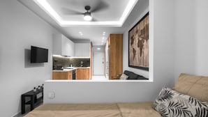 Ein modernes Schlafzimmer mit einem großen Bett, einem an der Wand montierten Fernseher, einer durch eine offene Tür sichtbaren Küche und einem Deckenventilator.