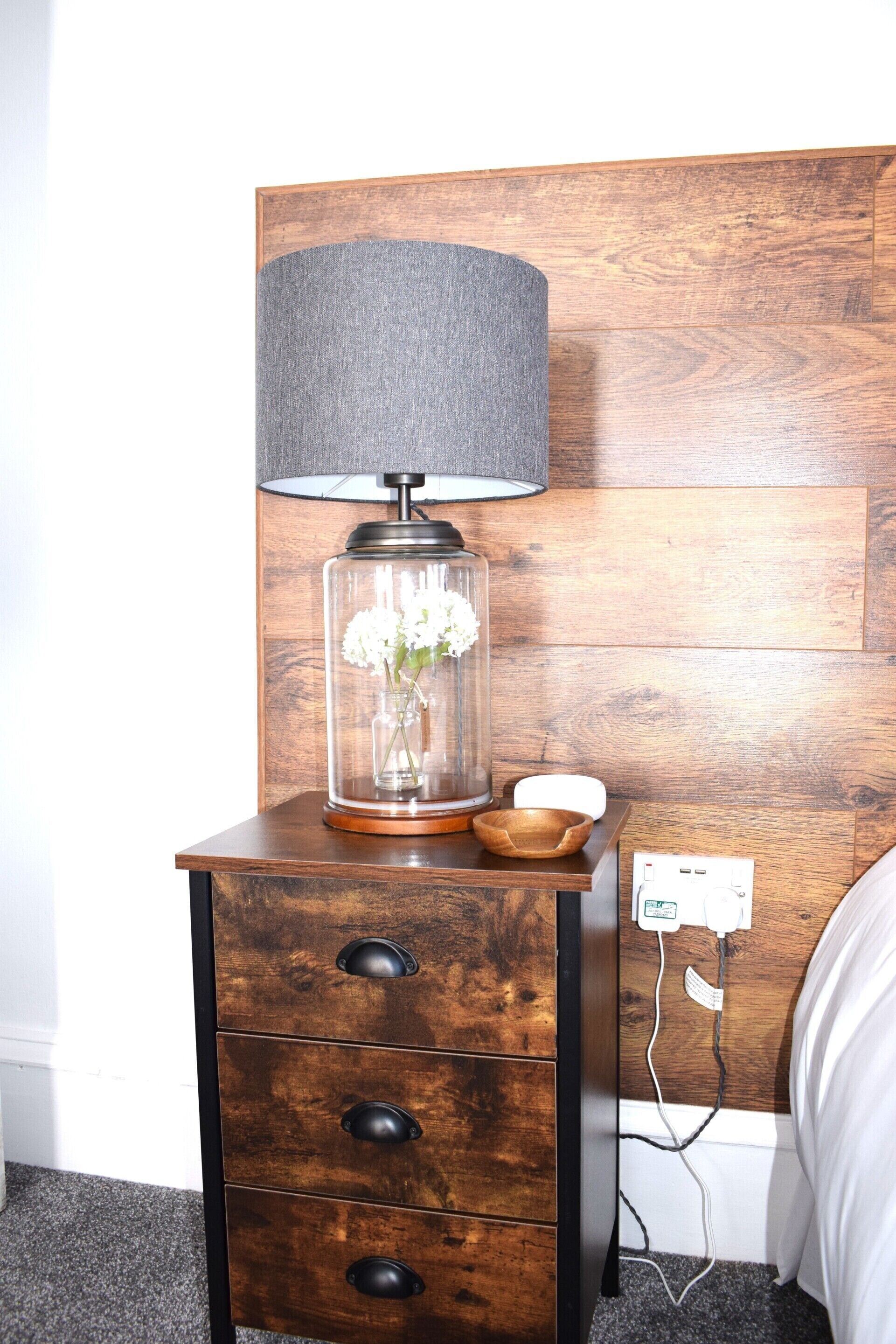 Une table de chevet en bois, une lampe, un vase avec des fleurs et une prise électrique.