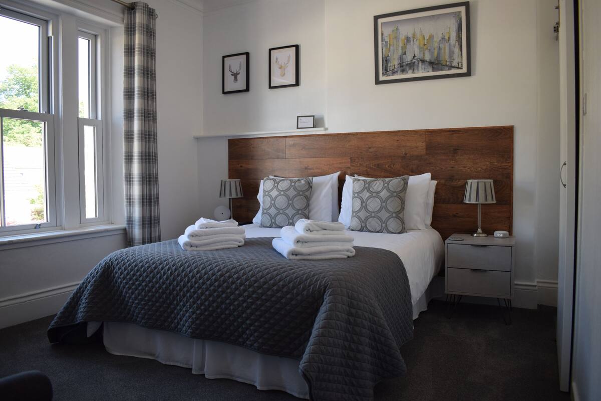A neatly made bed with a grey quilt, two bedside tables with lamps, and a cityscape painting on the wall.