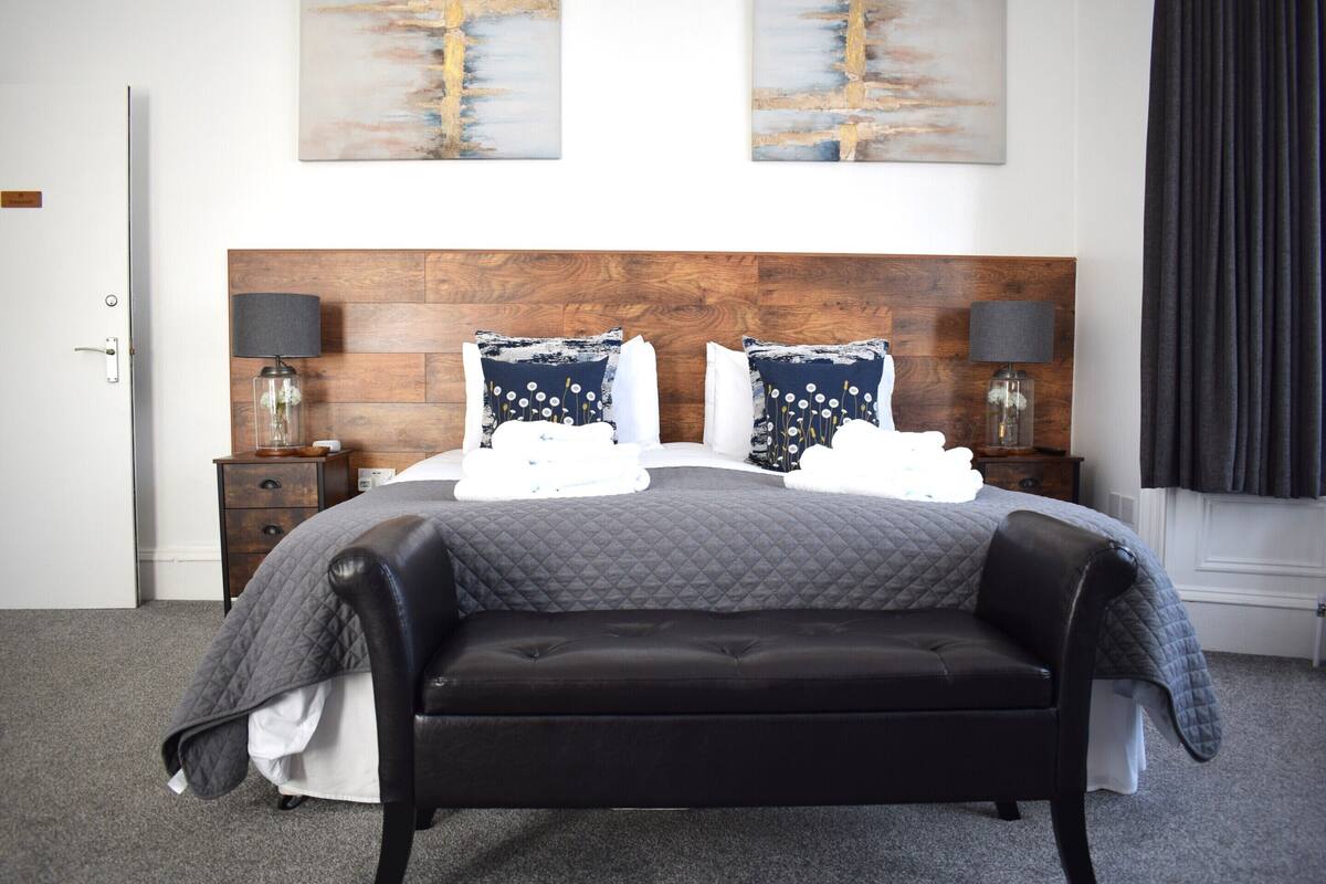 A bedroom with a wooden headboard, a black leather sofa, a bed with a quilted grey duvet, two bedside lamps, and two abstract paintings on the wall.