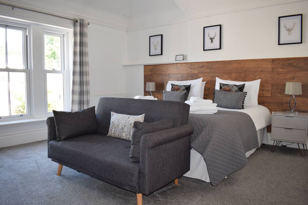 A modern bedroom with a grey sofa, a large bed with multiple pillows, a wooden headboard, and framed pictures on the wall.