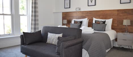 A modern bedroom with a grey sofa, a large bed with multiple pillows, a wooden headboard, and framed pictures on the wall.