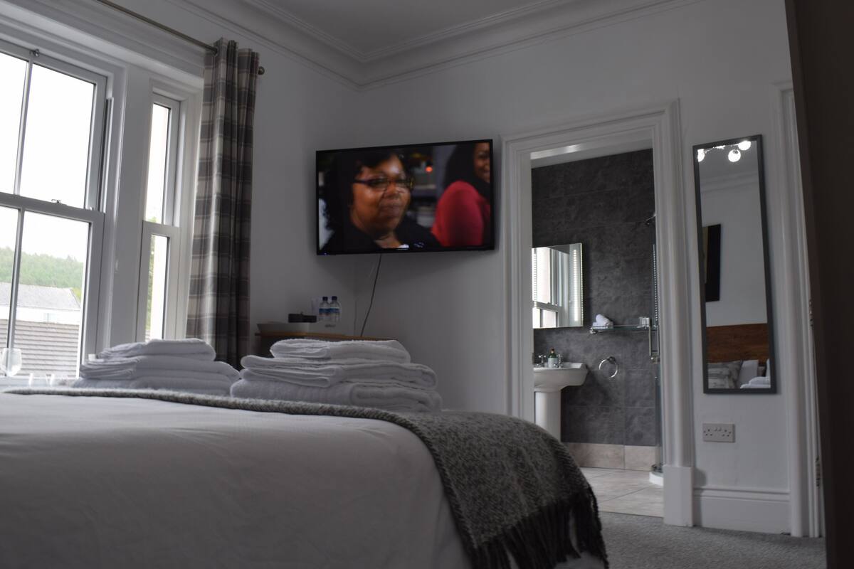 A hotel room with a large bed, a television mounted on the wall, a bathroom with a sink and mirror, and a view of the outdoors through the window.