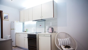 A compact kitchen with white cabinets, a stove, a refrigerator, and a coffee maker. There is a wicker chair with a pillow. - Downtown Penthouse Thessaloniki (Thessaloniki)