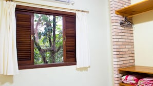 A room with a window featuring wooden shutters, a white curtain, and a view of trees outside. There is a brick wall and a shelf with folded blankets. - Pousada A Cor do Sol (Ubatuba)
