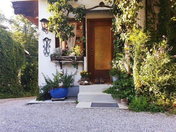 Una casa con puerta de madera, un pequeño porche con una maceta azul y plantas en macetas, y un camino de entrada de grava.