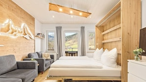 A modern bedroom with a wooden ceiling, a grey sofa, a bed with white bedding, and a view of mountains through large windows. - Ferienwohnungen Marienblick (Berchtesgaden)
