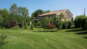 A rustic stone house with a tiled roof, surrounded by a well-maintained lawn and various shrubs.