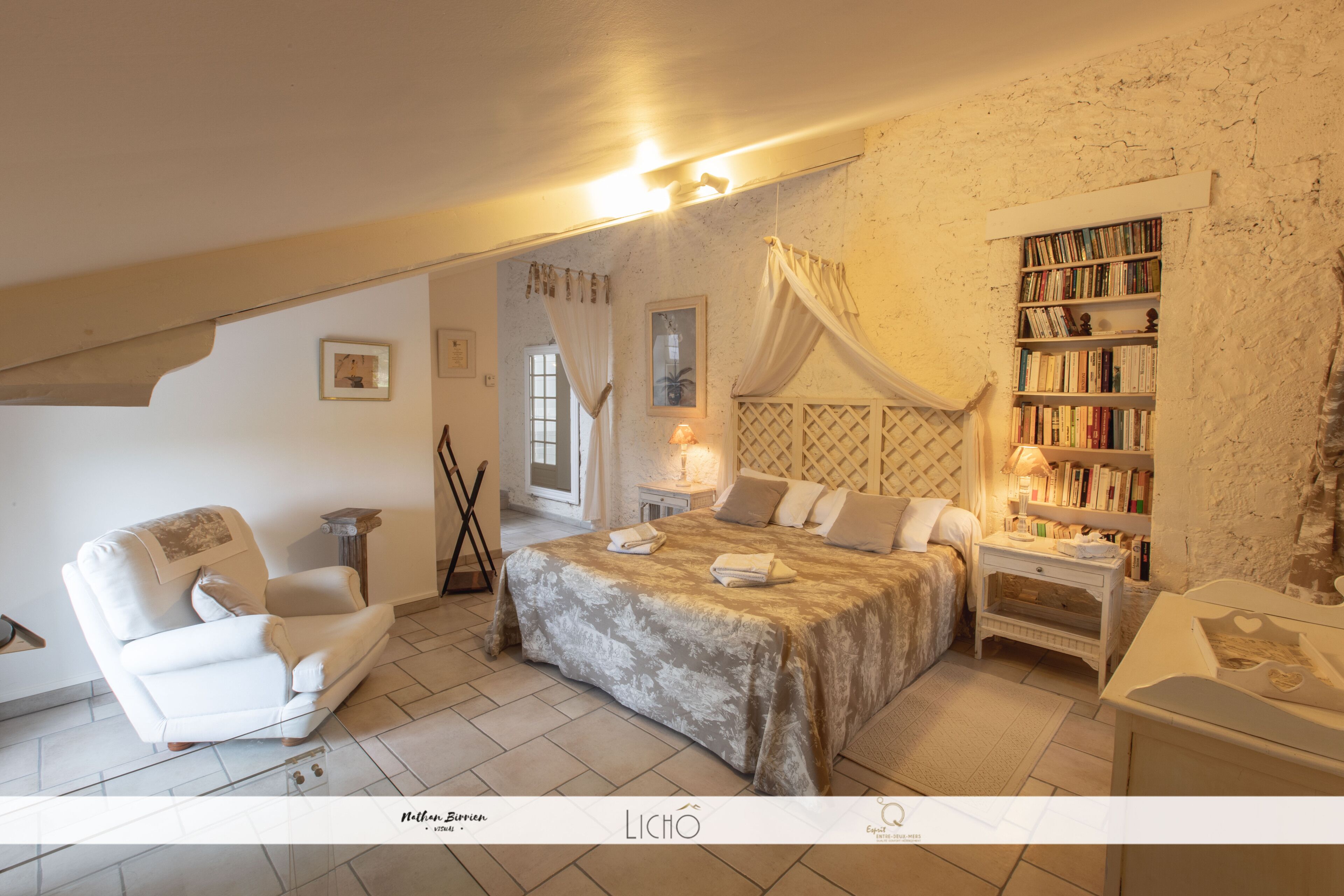 Een slaapkamer met een groot bed, een witte fauteuil, een nachtkastje met een lamp, een boekenkast en een raam met gordijnen.