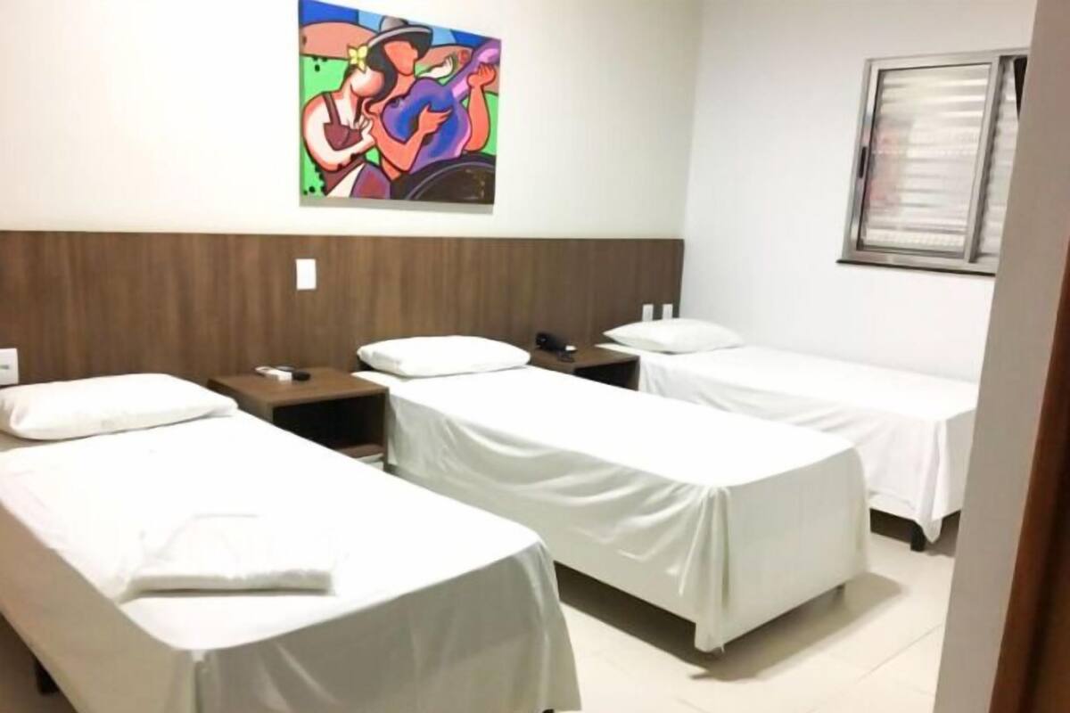 Habitación de hotel con dos camas individuales, cabecero de madera, mesita de noche con lámpara, un cuadro en la pared y una ventana con persianas.