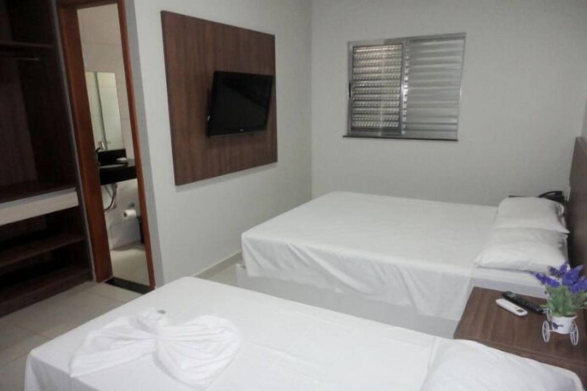 Habitación de hotel con dos camas individuales, un televisor montado en la pared y una ventana con persianas.