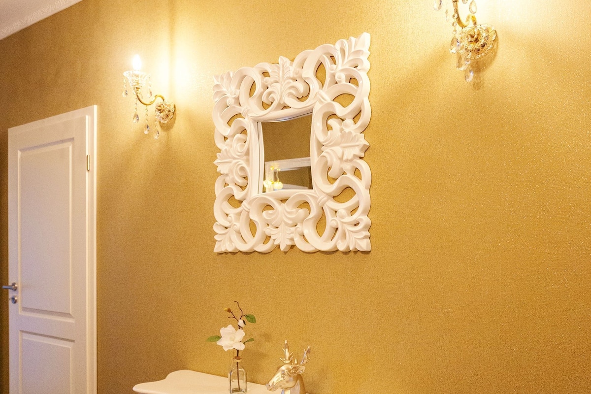Un couloir avec un mur doré, un grand miroir blanc orné et une console blanche sur laquelle sont posés un vase et une petite statue.